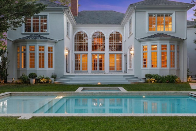 back house at dusk featuring a pool with hot tub and a lawn