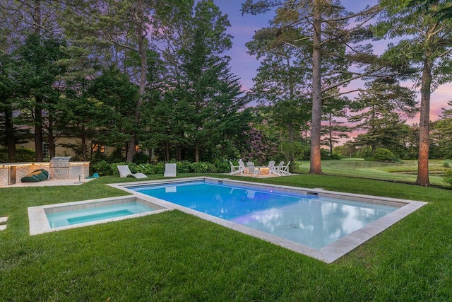 pool at dusk with an outdoor kitchen, grilling area, a lawn, and a fire pit