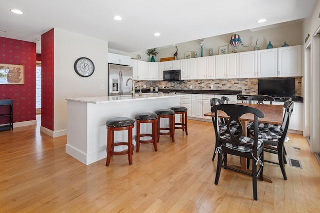 kitchen with stainless steel fridge, an island with sink, white cabinets, light hardwood / wood-style floors, and backsplash