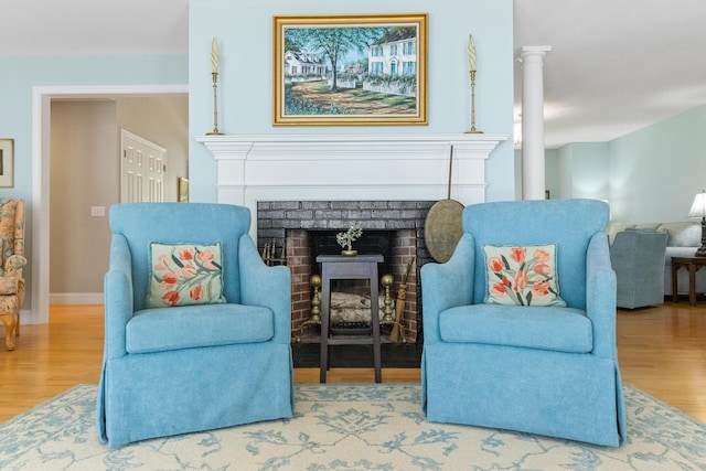 living area featuring baseboards, a brick fireplace, wood finished floors, and ornate columns