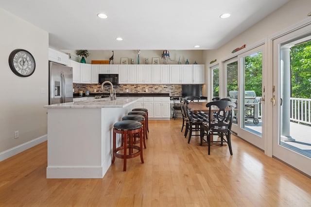 kitchen featuring white cabinets, backsplash, stainless steel fridge with ice dispenser, light stone countertops, and a center island with sink