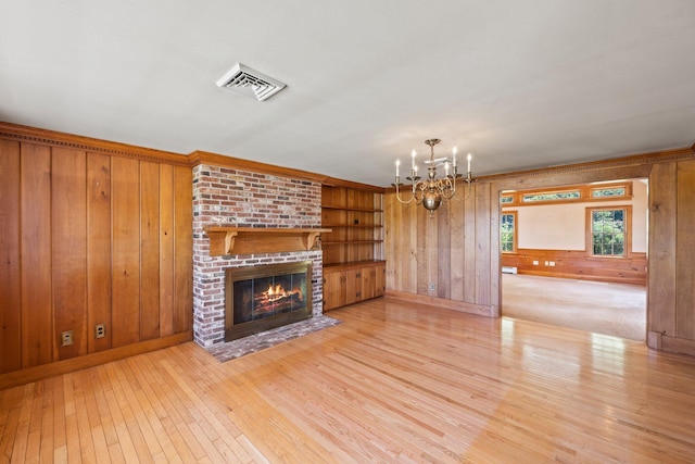 unfurnished living room featuring light hardwood / wood-style floors, a brick fireplace, built in shelves, an inviting chandelier, and wood walls