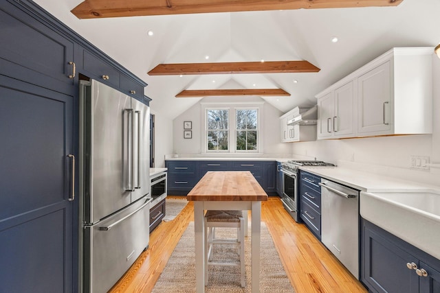 kitchen with blue cabinets, wall chimney exhaust hood, vaulted ceiling with beams, premium appliances, and light hardwood / wood-style floors