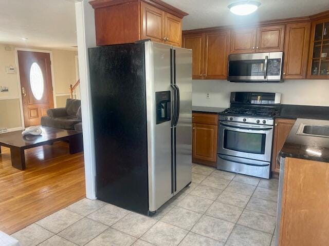 kitchen with glass insert cabinets, brown cabinetry, light tile patterned floors, appliances with stainless steel finishes, and dark countertops