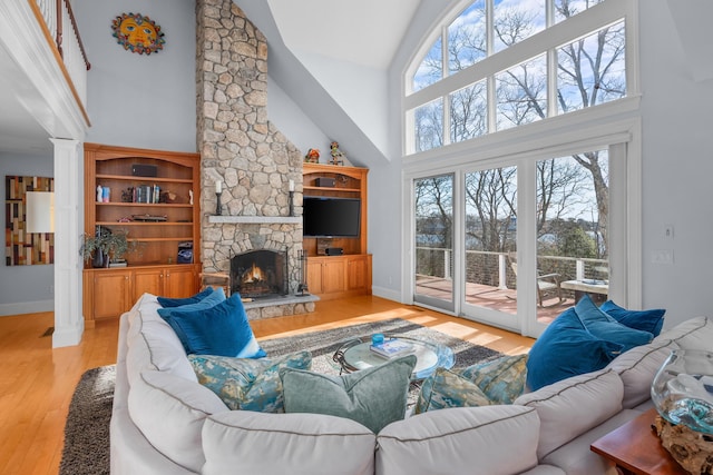 living room featuring ornate columns, light wood-style floors, a stone fireplace, high vaulted ceiling, and baseboards