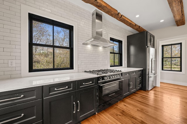 kitchen featuring premium appliances, wall chimney range hood, dark cabinetry, beamed ceiling, and light wood-style floors