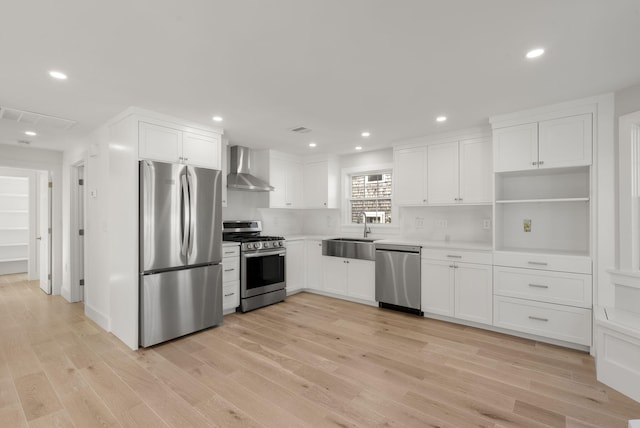 kitchen with light wood finished floors, stainless steel appliances, white cabinets, a sink, and wall chimney exhaust hood