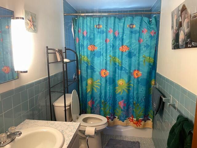 bathroom with tile walls, a wainscoted wall, toilet, shower / bath combo with shower curtain, and vanity