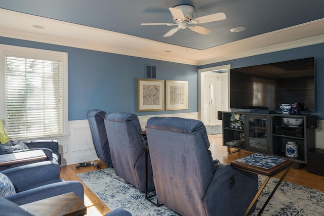 cinema room with wood finished floors, visible vents, ceiling fan, ornamental molding, and wainscoting