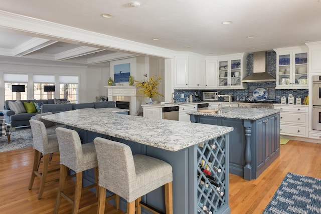 kitchen with wall chimney range hood, an island with sink, light wood-style flooring, and white cabinets