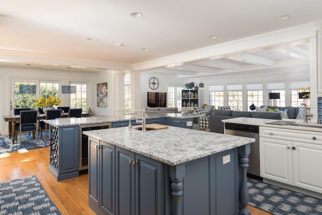 kitchen with an island with sink, a sink, stainless steel dishwasher, light wood-style floors, and ornate columns