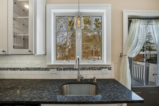 kitchen with a sink, backsplash, dark stone counters, glass insert cabinets, and pendant lighting