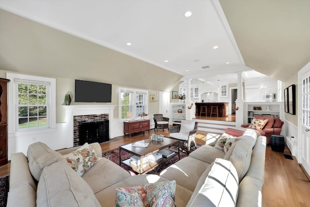 living room with light hardwood / wood-style floors, ornamental molding, vaulted ceiling, a brick fireplace, and decorative columns