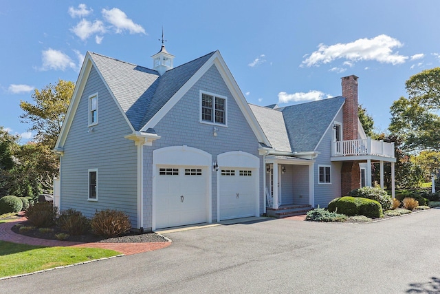 view of front of property featuring asphalt driveway, a garage, a shingled roof, a balcony, and a chimney