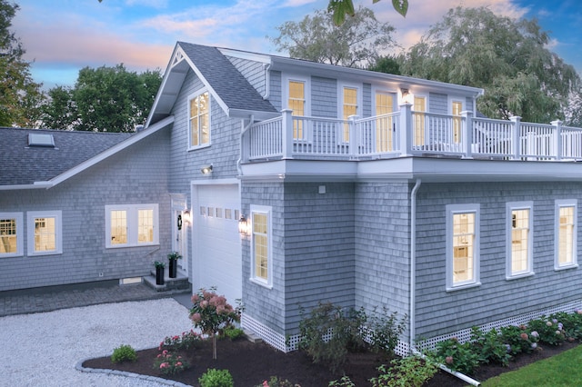 back of house at dusk featuring a shingled roof, a balcony, and aphalt driveway