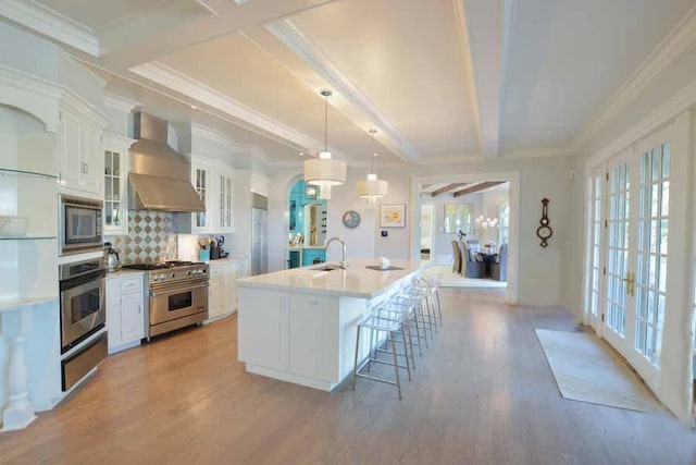 kitchen with decorative light fixtures, white cabinetry, an island with sink, stainless steel appliances, and wall chimney exhaust hood