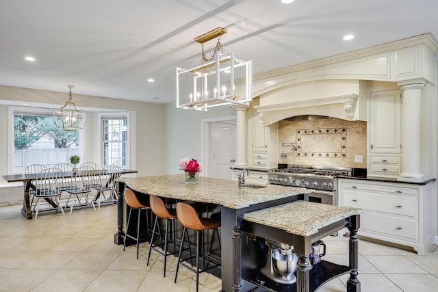 kitchen featuring stainless steel stove, a kitchen island with sink, dark stone countertops, decorative light fixtures, and backsplash