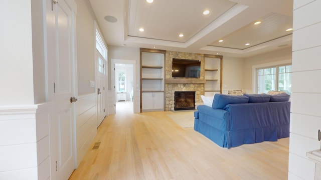 living room featuring crown molding, built in features, a stone fireplace, a tray ceiling, and light hardwood / wood-style floors