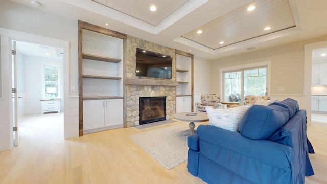 living room with built in shelves, a fireplace, light wood-type flooring, a tray ceiling, and crown molding