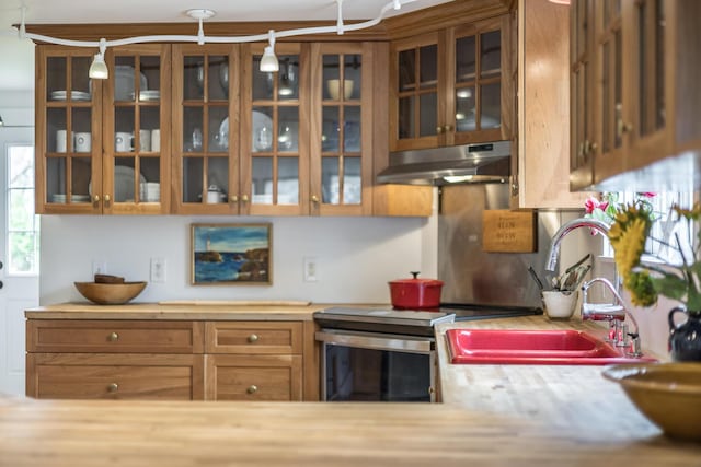 kitchen with under cabinet range hood, glass insert cabinets, stainless steel range with electric cooktop, a sink, and wood counters