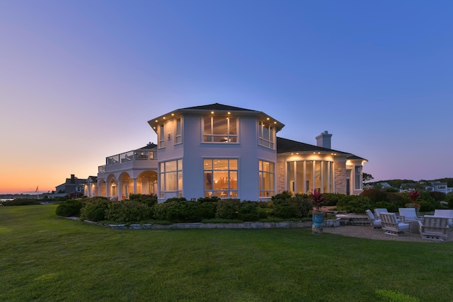 back house at dusk featuring a balcony, a patio area, and a lawn