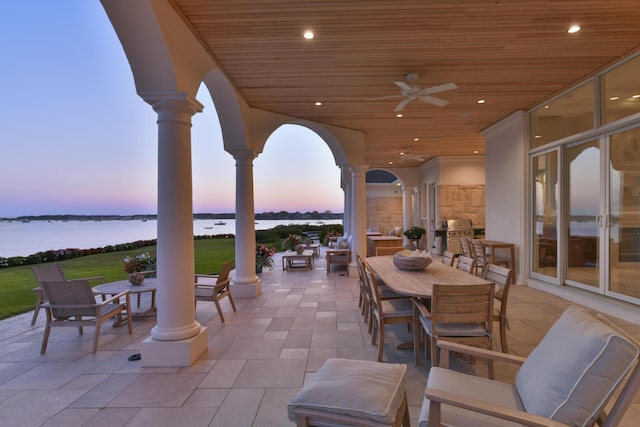 patio terrace at dusk with ceiling fan, a water view, exterior kitchen, and a grill