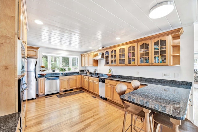 kitchen featuring wall chimney range hood, a breakfast bar area, stainless steel appliances, light hardwood / wood-style floors, and dark stone counters