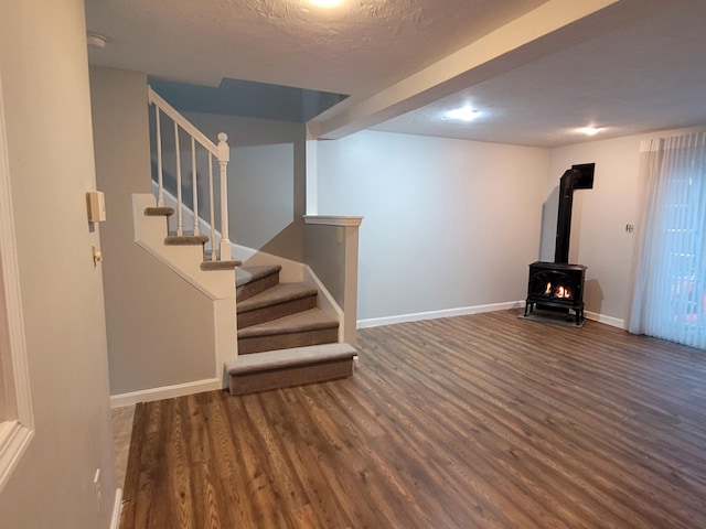 below grade area featuring a wood stove, a textured ceiling, baseboards, wood finished floors, and stairway