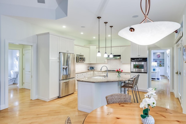 kitchen featuring decorative light fixtures, white cabinetry, light stone counters, stainless steel appliances, and a center island with sink