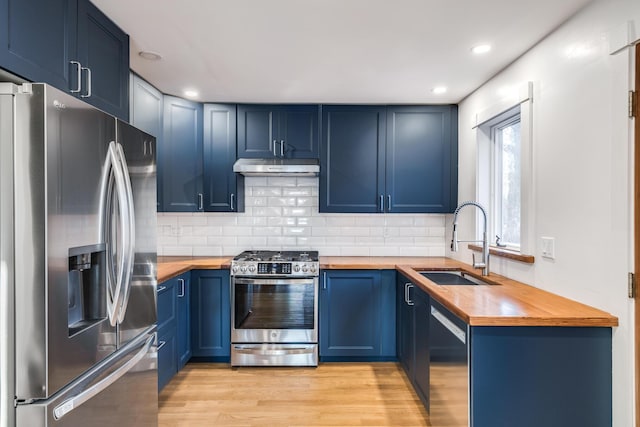 kitchen with under cabinet range hood, butcher block counters, a sink, blue cabinetry, and appliances with stainless steel finishes