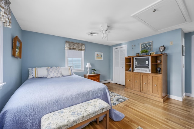 bedroom featuring baseboard heating, light wood-type flooring, a closet, attic access, and a ceiling fan