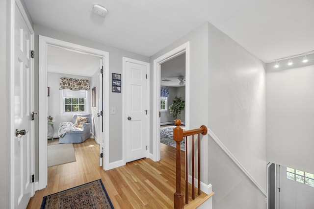 corridor with an upstairs landing, light wood finished floors, rail lighting, baseboards, and a baseboard radiator