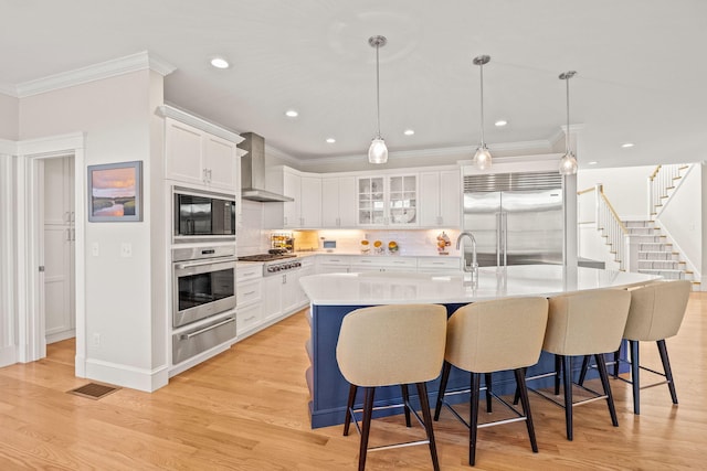 kitchen featuring white cabinetry, decorative light fixtures, built in appliances, a large island with sink, and wall chimney range hood