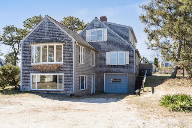 rear view of property featuring dirt driveway, an attached garage, a shingled roof, and a chimney