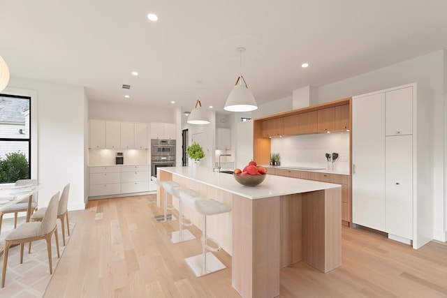 kitchen featuring white cabinetry, hanging light fixtures, light wood-type flooring, a center island with sink, and double oven