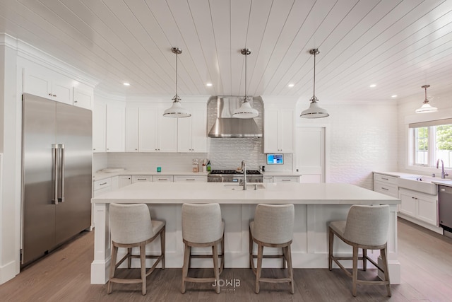 kitchen featuring pendant lighting, wall chimney range hood, appliances with stainless steel finishes, and an island with sink