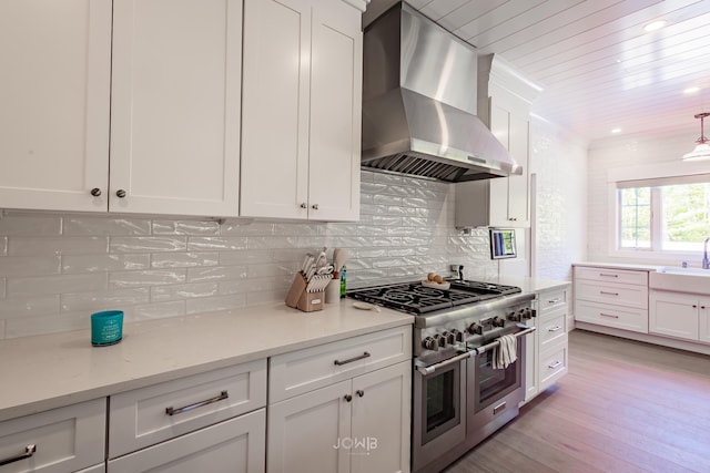 kitchen with light stone countertops, white cabinetry, wall chimney range hood, sink, and range with two ovens