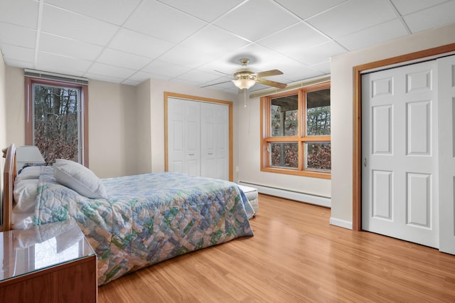 bedroom featuring two closets, a paneled ceiling, wood-type flooring, a baseboard heating unit, and ceiling fan