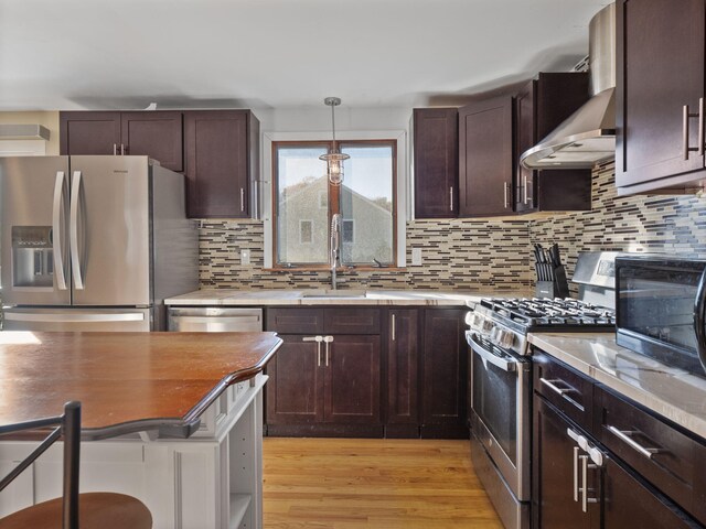 kitchen featuring appliances with stainless steel finishes, decorative backsplash, decorative light fixtures, light hardwood / wood-style flooring, and wall chimney exhaust hood