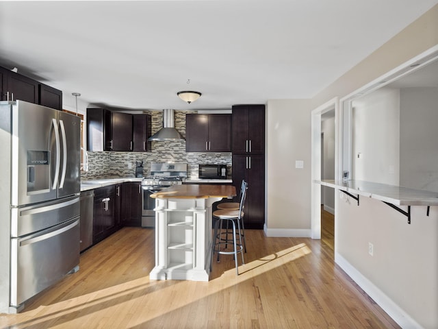 kitchen featuring light hardwood / wood-style flooring, appliances with stainless steel finishes, wall chimney exhaust hood, decorative backsplash, and a breakfast bar area