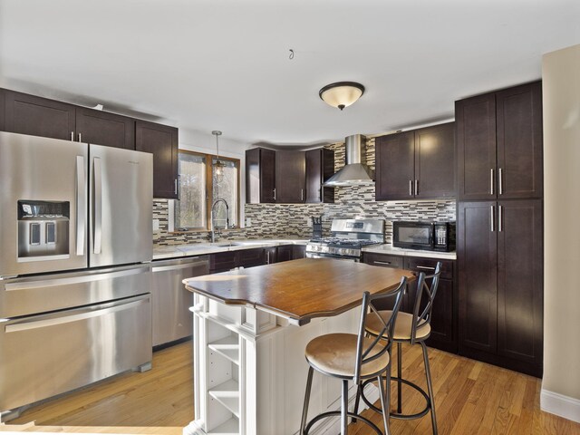 kitchen featuring pendant lighting, appliances with stainless steel finishes, a kitchen island, wall chimney range hood, and light wood-type flooring