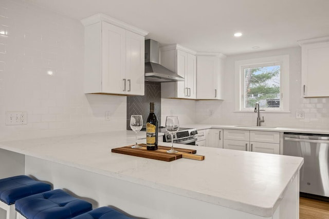 kitchen with a peninsula, wall chimney exhaust hood, white cabinetry, a sink, and appliances with stainless steel finishes