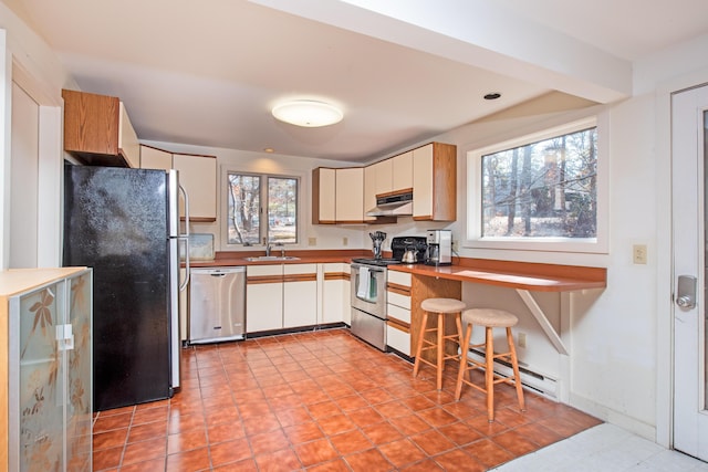 kitchen featuring light tile patterned floors, a kitchen breakfast bar, stainless steel appliances, under cabinet range hood, and a sink