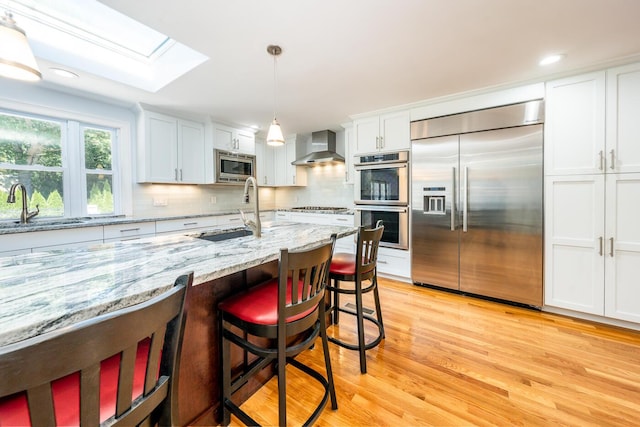 kitchen featuring built in appliances, decorative light fixtures, wall chimney range hood, and white cabinets