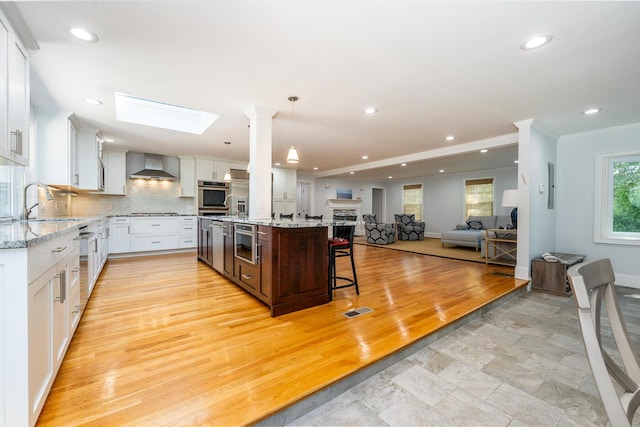 kitchen featuring stainless steel oven, white cabinetry, wall chimney range hood, a center island with sink, and a breakfast bar area