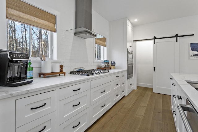 kitchen featuring appliances with stainless steel finishes, white cabinetry, light stone countertops, a barn door, and wall chimney exhaust hood