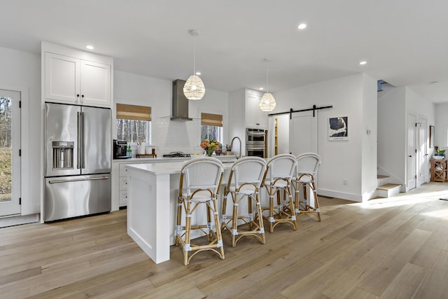 kitchen featuring a center island with sink, wall chimney range hood, stainless steel appliances, a barn door, and white cabinets