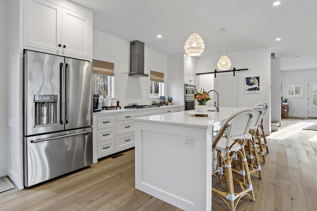 kitchen featuring light stone counters, stainless steel appliances, a barn door, wall chimney range hood, and light wood-type flooring