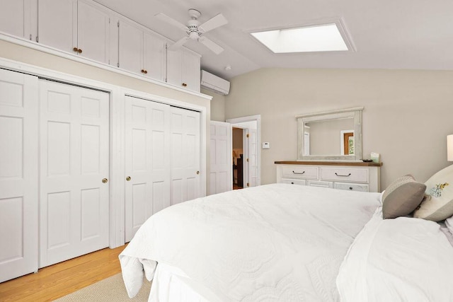 bedroom with lofted ceiling with skylight, two closets, light wood-type flooring, ceiling fan, and a wall unit AC