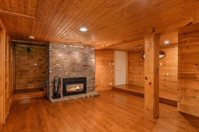 unfurnished living room with light wood-type flooring, a brick fireplace, track lighting, wooden ceiling, and wood walls